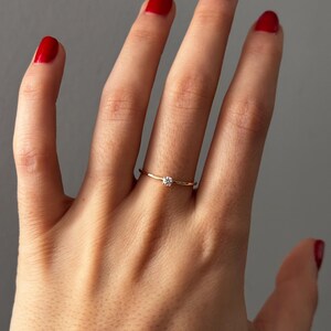 May include: A delicate gold ring with a small, round, clear gemstone. The ring is worn on a finger with red nail polish. The ring's band is thin and simple, highlighting the single gemstone.
