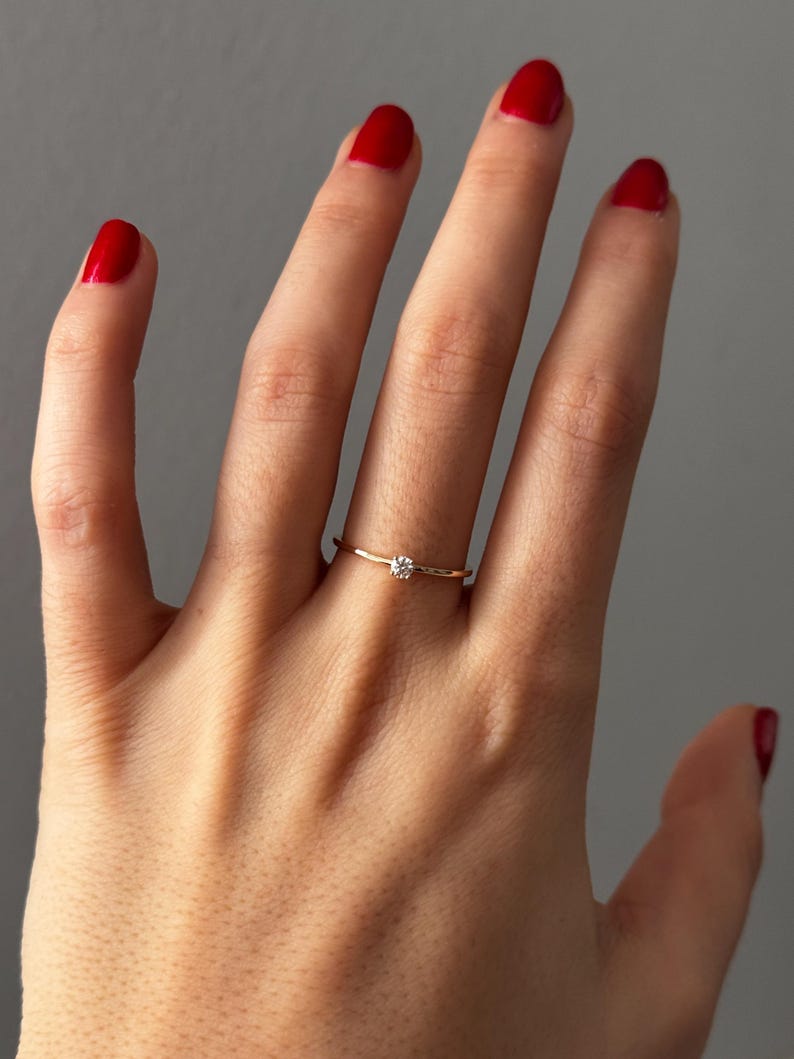 May include: A delicate gold ring with a small, round, clear gemstone. The ring is worn on a finger with red nail polish. The ring's band is thin and simple, highlighting the single gemstone.