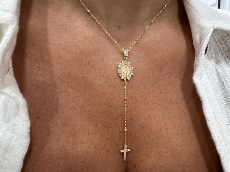 May include: Gold-toned necklace featuring a pendant with a religious motif, surrounded by sparkling accents. The necklace has a delicate chain with a cross pendant at the end. The design is elegant and suitable for religious occasions.