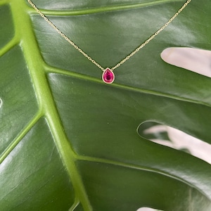 May include: A delicate gold chain necklace with a single teardrop-shaped red gemstone pendant.