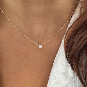 May include: A fine gold necklace featuring a single, clear, round gemstone pendant. The necklace is worn against skin, with a white shirt in the background. The design is minimalist, highlighting the pendant's sparkle.