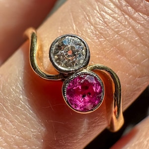 Könnte beinhalten: Ein goldener Ring mit einem einzigartigen Design, der zwei runde Edelsteine ​​zeigt. Einer ist ein klarer Diamant, der andere ein leuchtend roter Rubin. Der Ring hat eine geschwungene Form, die ein elegantes Schmuckstück ergibt.