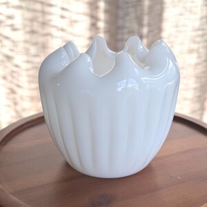 May include: A white, ruffled-edge glass bowl sits on a round, wooden tray. The bowl has a textured, ribbed design and a glossy finish. The wooden tray is a warm brown color and has a smooth surface. The background is a soft, neutral color.