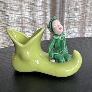 May include: A vintage ceramic figurine featuring a green elf seated on a lime green shoe-shaped planter. The elf has a pink face, red lips, and is wearing a green outfit. The planter has a glossy finish.