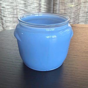 May include: A pale blue ceramic jar with a clear glass rim and two small handles. The jar is on a dark wooden surface. The background shows a sheer, light-coloured curtain.