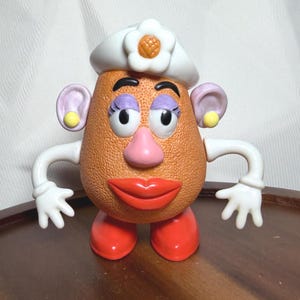 May include: A vintage Mrs. Potato Head toy with a textured orange body, large red lips, and a pink nose. She wears a white hat with a flower and has purple eyelashes, yellow earrings, and white arms and hands. Her red feet are visible.