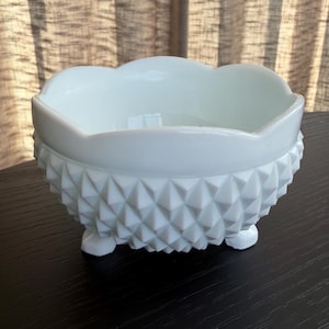May include: A white, scalloped-edge bowl with a textured, geometric pattern around the sides. The bowl sits on three small feet. The bowl is made of a solid, opaque material.