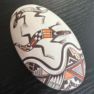May include: Oval-shaped ceramic art piece with a cream-colored base. Two stylized lizard figures in black, orange, and red are depicted. The bottom features geometric patterns in brown, orange, and green. A unique decorative item.