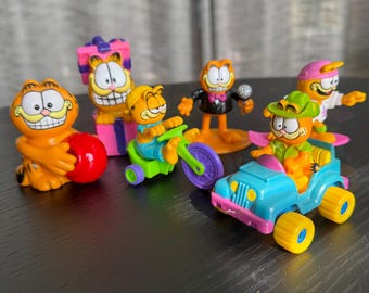 May include: A collection of Garfield figurines in various poses. One figurine is holding a red ball, while others are riding a motorcycle, a jeep, and a surfboard. Another is standing with a microphone, and one is next to a gift box.