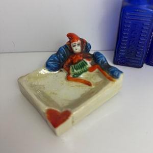 May include: A vintage ceramic ashtray with a reclining jester figure in red, blue, and green. The ashtray is rectangular with a red heart on one corner. Two cobalt blue glass bottles are in the background.