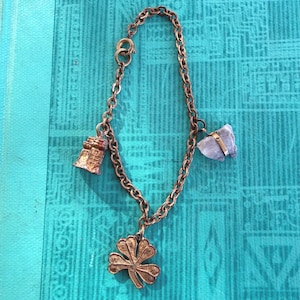 May include: A vintage charm bracelet with a bronze chain and three charms. Charms include a bell, a four-leaf clover, and a light purple stone. The bracelet is displayed on a turquoise patterned surface.
