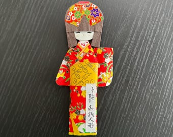 May include: A colorful origami doll in a traditional Japanese kimono. The doll features a red kimono with floral patterns, a yellow obi, and a decorative headdress. The doll has a smiling face and long brown hair. Japanese text is visible on the doll.