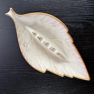 May include: A leaf-shaped ashtray with a white base and gold trim. The ashtray has a textured surface with gold accents and a series of small, rectangular cigarette rests. The design is reminiscent of a stylized leaf.