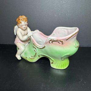 May include: A vintage ceramic shoe-shaped planter with a cherub figurine. The planter is painted in shades of pink and green with gold accents. The cherub has blonde hair and is wearing a pink garment.