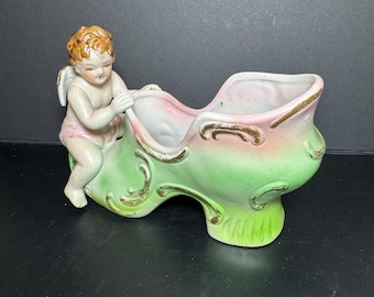 May include: A vintage ceramic shoe-shaped planter with a cherub figurine. The planter is painted in shades of pink and green with gold accents. The cherub has blonde hair and is wearing a pink garment.