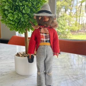 May include: A vintage doll wearing a red jacket, grey pants, a grey hat, and a plaid shirt. The doll is holding a small black bucket.