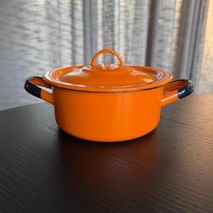 May include: A bright orange enameled metal pot with a matching lid and two side handles. The pot features a smooth, glossy finish with dark blue accents on the handle tips and a thin dark blue rim around the top edge of the pot and the lid.