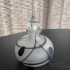 May include: A frosted glass perfume bottle with a clear, elongated stopper. The bottle has a round shape with black and purple abstract designs. The bottle is sitting on a black surface.