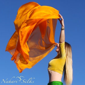 Può includere: Una donna danza con una sciarpa di seta arancione brillante contro un cielo blu. La sciarpa fluttua nel vento, creando un'immagine dinamica e colorata. La donna indossa un top corto giallo e una gonna verde.