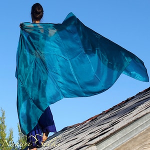 Op de afbeelding: Een vloeiende, turquoise zijden sjaal wordt omhoog gehouden tegen een heldere blauwe lucht. De sjaal is gedrapeerd en golvend, met zichtbare plooien en een glinsterende textuur. De foto is buiten genomen, met een dak op de voorgrond.