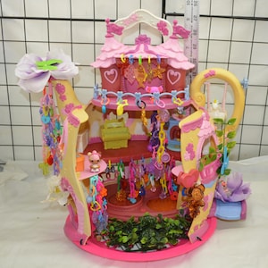 Op de afbeelding: Een roze en gele speelset in de vorm van een theepot, met een ontwerp met meerdere niveaus en hangende accessoires. De speelset bevat een schommel, decoratieve bloemen en een klein figuurtje. De hoogte van de speelset is ongeveer 30 cm.