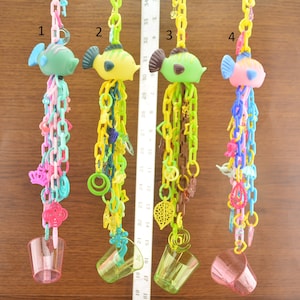 May include: Four colorful plastic bird toys with different colored fish on top and a cup at the bottom. The toys are hanging from a wooden surface.