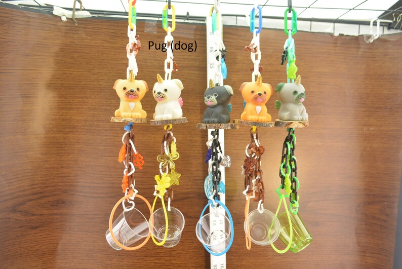 May include: A collection of colorful dog-themed hanging toys. Each toy features a pug figurine with a unicorn horn, suspended from a chain. The toys are attached to wooden platforms and adorned with various decorative elements, including plastic rings and chains.