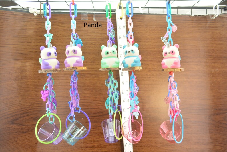 May include: A collection of colorful bird toys featuring panda-unicorn figures. Each toy has a panda figurine with a unicorn horn, wings, and a small wooden perch. The toys are suspended by chains and include hanging rings and cups.