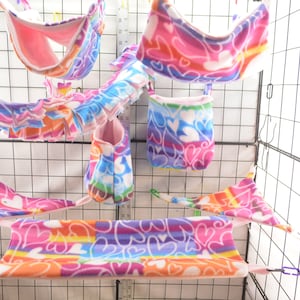 以下が含まれることがあります： 虹色とピンクのフリース素材のハムスターケージ用アクセサリーセット。アクセサリーには、ハンモック、吊り下げ式ポーチ、トンネルが含まれています。アクセサリーはすべてハート柄で飾られています。