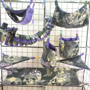 以下が含まれることがあります： 小型ペット用の紫と黒のパターン付きハンモック6個セット。ハンモックは柔らかなフリース製で、骸骨と花のデザインが施されています。