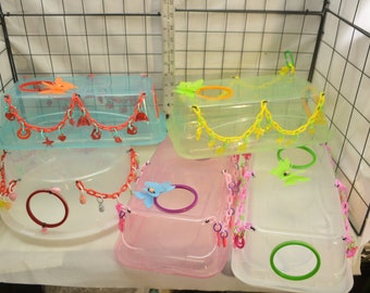 Puede incluir: Una colección de jaulas para mascotas de plástico transparente y colorido. Cada jaula tiene una tapa con bisagras y está adornada con cadenas y abalorios decorativos. Las jaulas vienen en varios colores, incluyendo azul, rosa y verde, y tienen aberturas circulares.