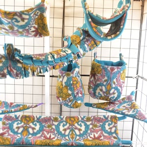 May include: A set of colourful hanging pet beds and toys for small animals. The beds and toys are made of soft fabric with a floral pattern and blue trim. The set includes a hammock, a tunnel, a couple of pouches, and a hanging bed.