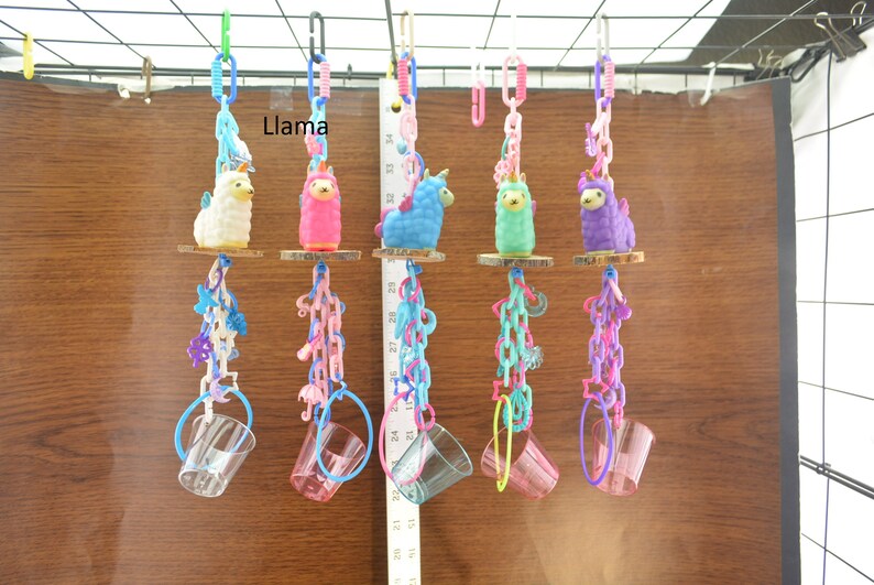 May include: A collection of colorful llama-themed bird toys. Each toy features a small llama figure in various colors, suspended by chains and a clear plastic cup. The word "Llama" is visible in the image.