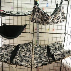 Puede incluir: Una colección de accesorios para mascotas pequeñas, que incluyen hamacas y ropa de cama, en un patrón de calaveras y símbolos ocultos en blanco y negro. Los artículos están suspendidos dentro de una jaula de alambre, mostrando una variedad de formas y tamaños.