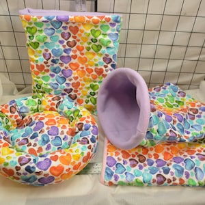 Puede incluir: Un conjunto de tres camas para mascotas con un estampado de corazones de acuarela coloridos. Las camas incluyen una cama redonda, una cama rectangular y una cama colgante.