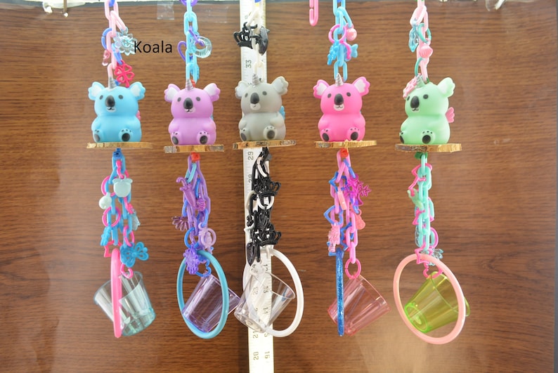 May include: Five colorful koala bear toys with unicorn horns hanging from chains. Each koala is a different color: blue, purple, gray, pink, and green. Each koala is attached to a small wooden disc and a clear plastic cup.