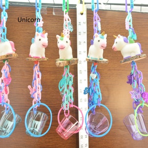 May include: A collection of five unicorn-themed bird toys. Each toy features a white unicorn figurine atop a wooden perch, connected by colorful plastic chains and rings. The chains are in shades of pink, blue, and purple. The word "Unicorn" is visible.