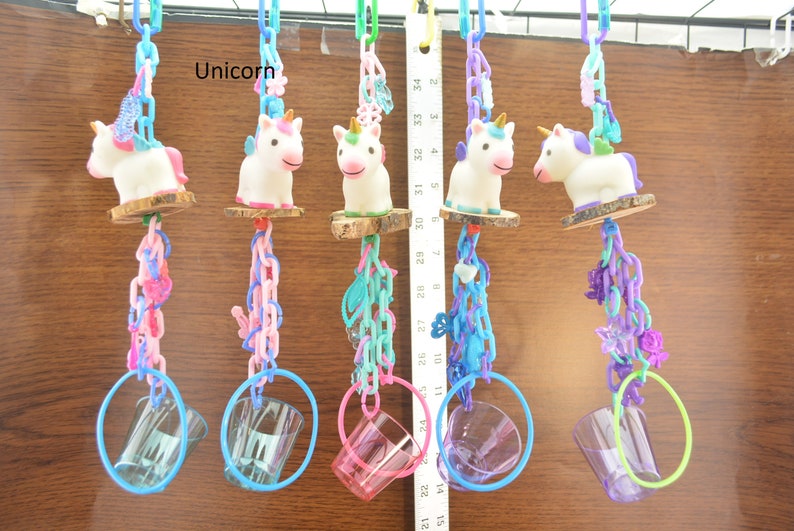 May include: A collection of five unicorn-themed bird toys. Each toy features a white unicorn figurine atop a wooden perch, connected by colorful plastic chains and rings. The chains are in shades of pink, blue, and purple. The word "Unicorn" is visible.