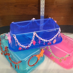 May include: Three colorful plastic containers, blue, teal, and pink, are stacked. Each container has a lid and is decorated with chains and charms. The containers are rectangular and appear to be storage boxes.