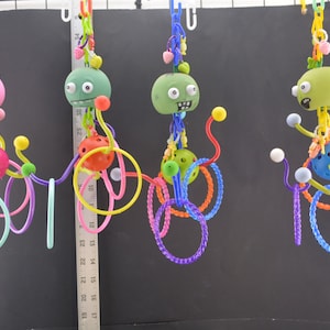 May include: Four colorful hanging toys for birds. Each toy features a green or pink head with googly eyes, a ball, and multiple rings. The toys are made of plastic and are designed to be hung from a cage.