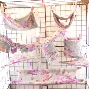 May include: A set of six hanging hamster cage accessories in a purple and grey butterfly print. The accessories include a hammock, a hanging pouch, a tunnel, and a hanging toy.