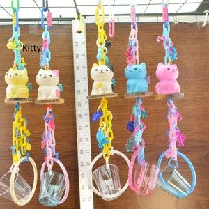May include: A collection of colorful cat-shaped toys with unicorn horns, wings, and attached clear plastic cups. The toys are suspended by chains and rings, with the word "Kitty" visible. The cats are yellow, white, blue, and pink.