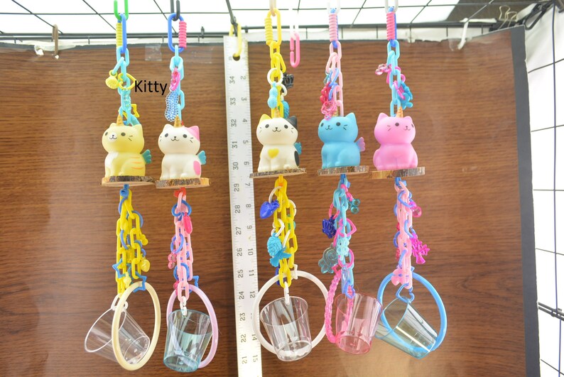 May include: A collection of colorful cat-shaped toys with unicorn horns, wings, and attached clear plastic cups. The toys are suspended by chains and rings, with the word "Kitty" visible. The cats are yellow, white, blue, and pink.