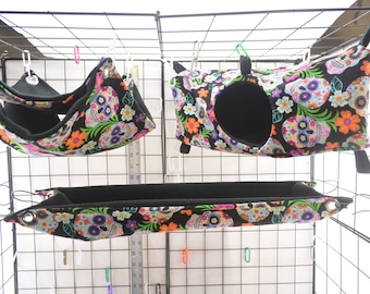 May include: Three hanging pet beds with a black and white skull and floral print. The beds are made of soft fabric and have a black fleece lining.