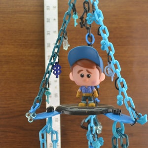 May include: A blue and black plastic toy hanging mobile with a small blue and white figurine of a boy wearing a blue hat and blue overalls. The mobile has multiple blue plastic chains with various charms.