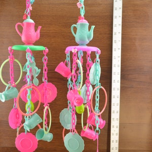 Op de afbeelding: Twee kleurrijke plastic vogel speeltjes met theepotten, kopjes, schoteltjes en ringen die aan kettingen hangen. De speeltjes zijn roze, groen en blauw.