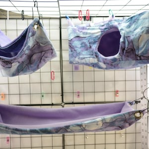 May include: Three purple and blue patterned hanging beds for small pets. The beds are made of soft fabric and have a variety of shapes, including a hammock, a tunnel, and a pouch.