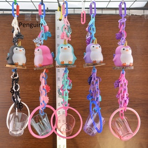 May include: A collection of colorful penguin-themed toys, each suspended by chains and rings. The penguins are in various colors, including black, pink, blue, and purple, and have golden horns. The toys are hanging against a wooden background.