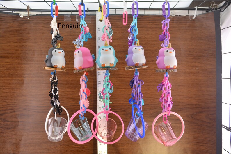 May include: A collection of colorful penguin-themed toys, each suspended by chains and rings. The penguins are in various colors, including black, pink, blue, and purple, and have golden horns. The toys are hanging against a wooden background.