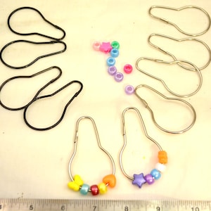 May include: A collection of shower curtain hooks and decorative beads. The hooks are black and silver, with some adorned with colorful beads in various shapes. The beads include stars, hearts, and round shapes in shades of yellow, orange, blue, and purple.